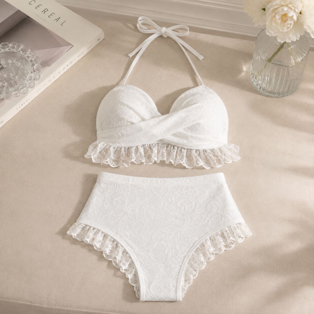 White two-piece bikini set with lace ruffle trim, laid flat on a beige fabric surface with decorative props including a book, glass bowl, and vase with flowers.