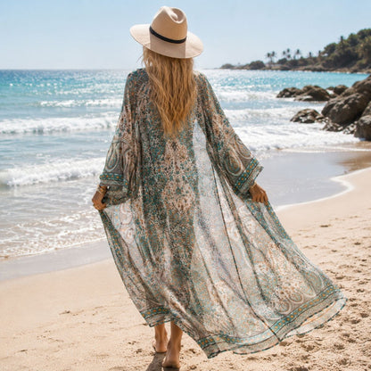 A woman viewed from behind on a sandy beach, wearing a long sheer bohemian-style maxi cover-up dress with a green, beige, and white paisley pattern, a wide-brimmed straw hat, and sandals.