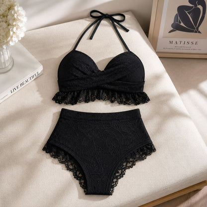 Black lace two-piece lingerie set consisting of a halter bralette and matching high-waisted briefs, arranged on a cream fabric surface with decorative props.