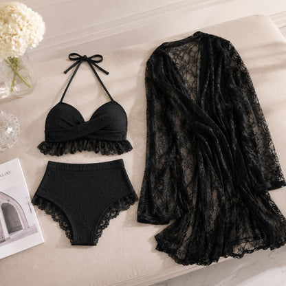 A three-piece women's swimwear set in black lace, including a halter bikini top, high-waisted bikini bottoms, and a sheer open-front cover-up, arranged on a beige upholstered surface with decorative props.