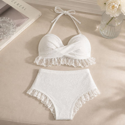 White two-piece bikini set with lace ruffle trim, laid flat on a beige fabric surface with decorative props including a book, glass bowl, and vase with flowers.