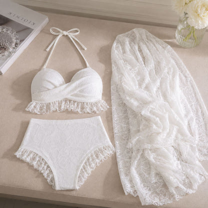 White lace lingerie set laid out on a beige surface, including a halter bra top, matching high-waisted briefs, and a sheer lace shawl. A book and flowers are partially visible as props.