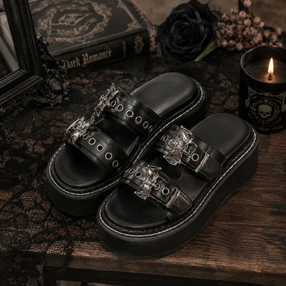 A pair of black platform slide sandals with ornate silver gothic-style hardware, displayed on a dark wooden table with a black lace cloth, a book, a black rose, and a lit candle.