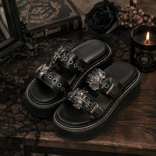 A pair of black platform slide sandals with ornate silver gothic-style hardware, displayed on a dark wooden table with a black lace cloth, a book, a black rose, and a lit candle.