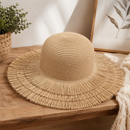 A natural straw sun hat with a wide frayed brim, displayed on a wooden table in a styled home setting with a potted plant and framed botanical print in the background.