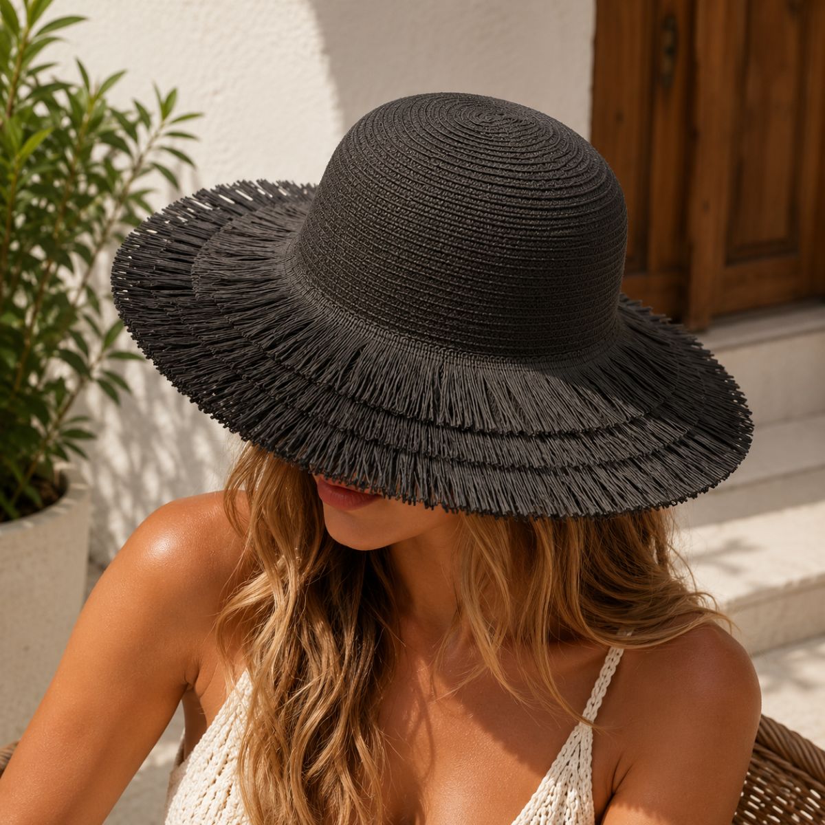 A woman wearing a wide-brimmed black woven sun hat with a frayed brim, seated outdoors in bright sunlight.