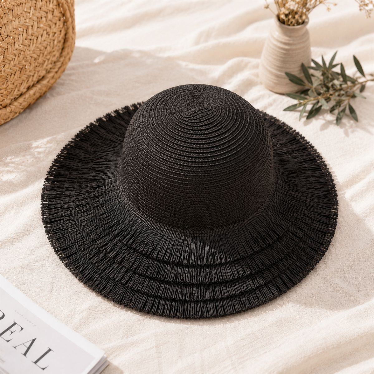 A black woven sun hat with a wide frayed brim, styled on a cream fabric surface with a straw basket edge, a small vase with dried flowers, and a book corner visible.