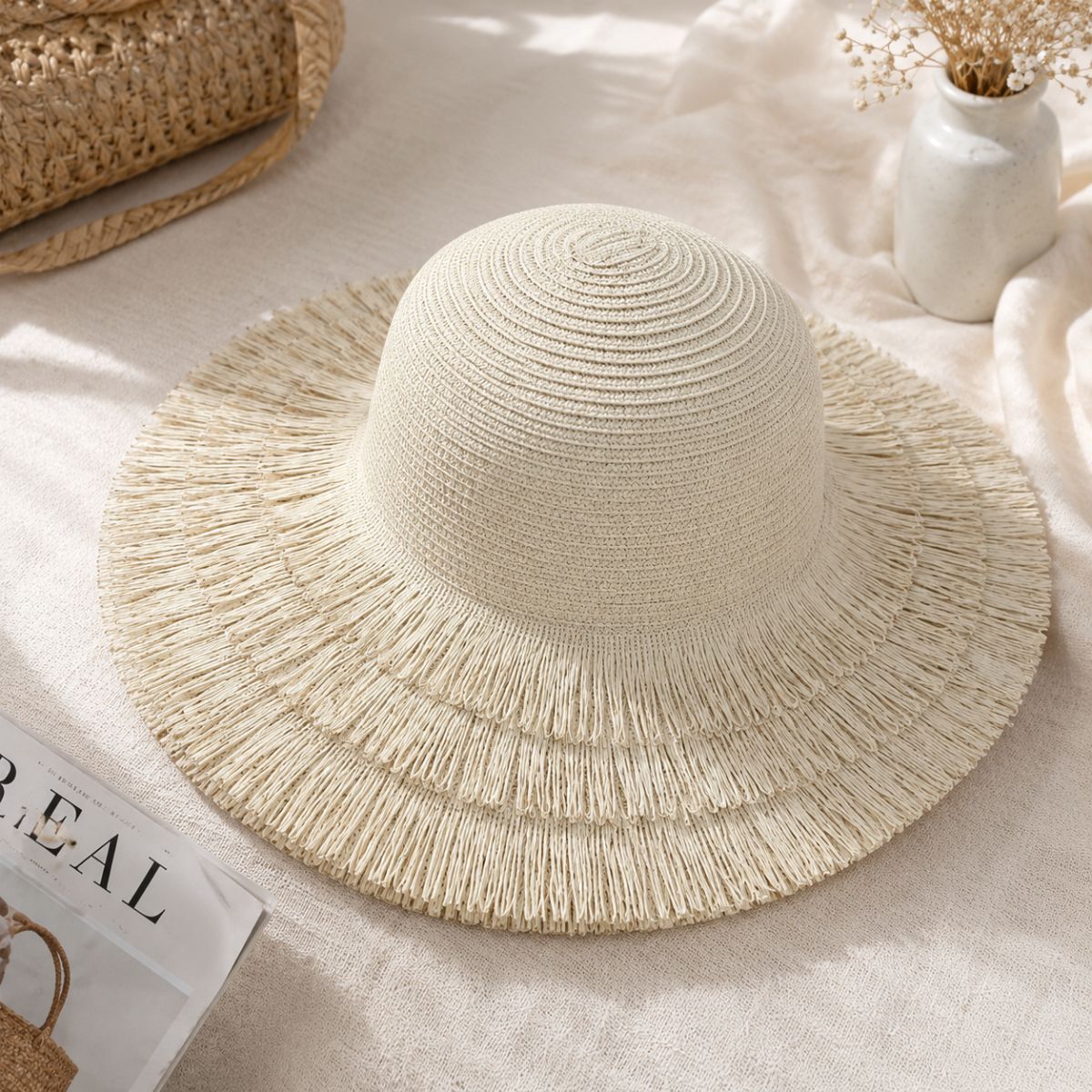 A wide-brimmed woven straw sun hat in a warm cream color, styled on a textured beige fabric surface with a white ceramic vase of dried flowers and a woven bag in the background.