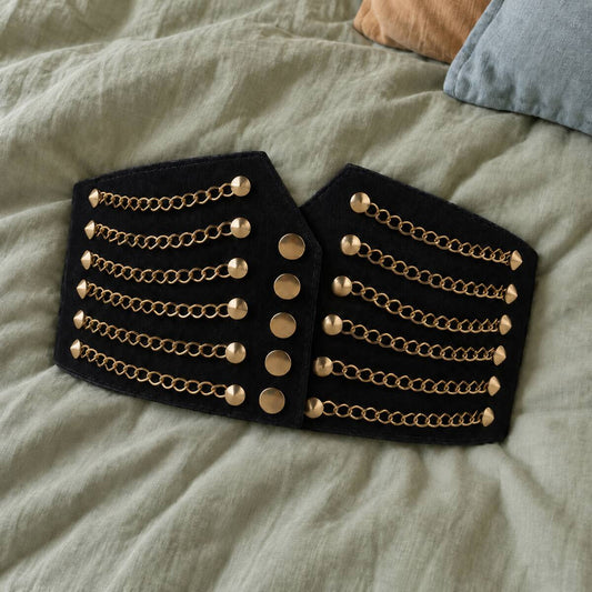Black elastic PU waist belt with gold-tone chains and studs resting on sage-green linen bedding.